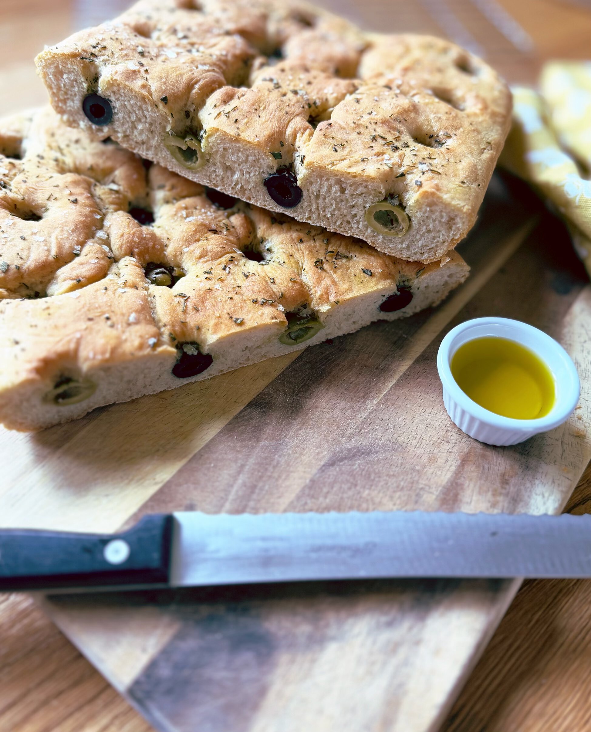 olive and rosemary focaccia on a wooden chopping board with a bread knife and pot of olive oil