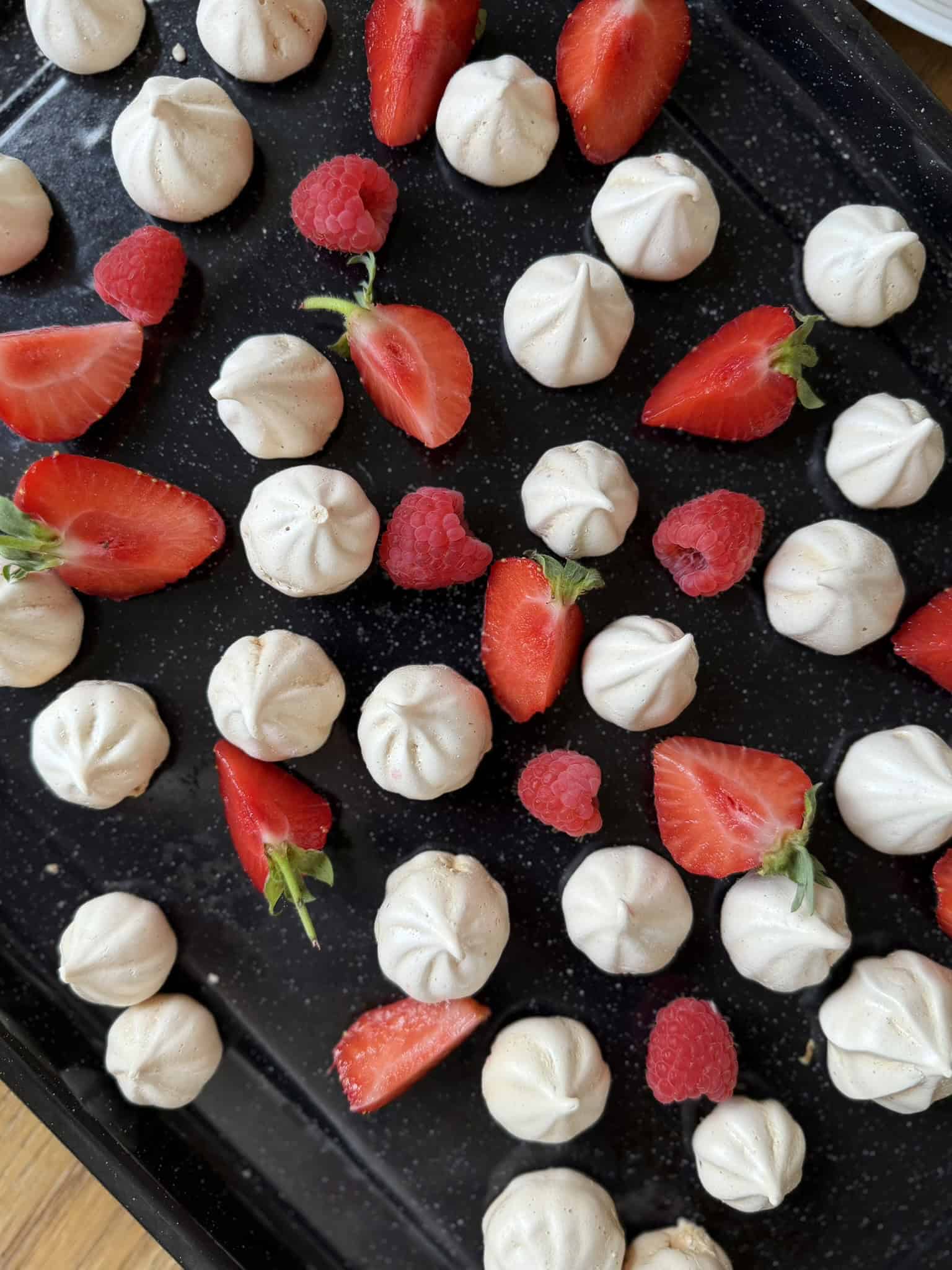 overhead shot of meringue kisses with red fruit on black tray