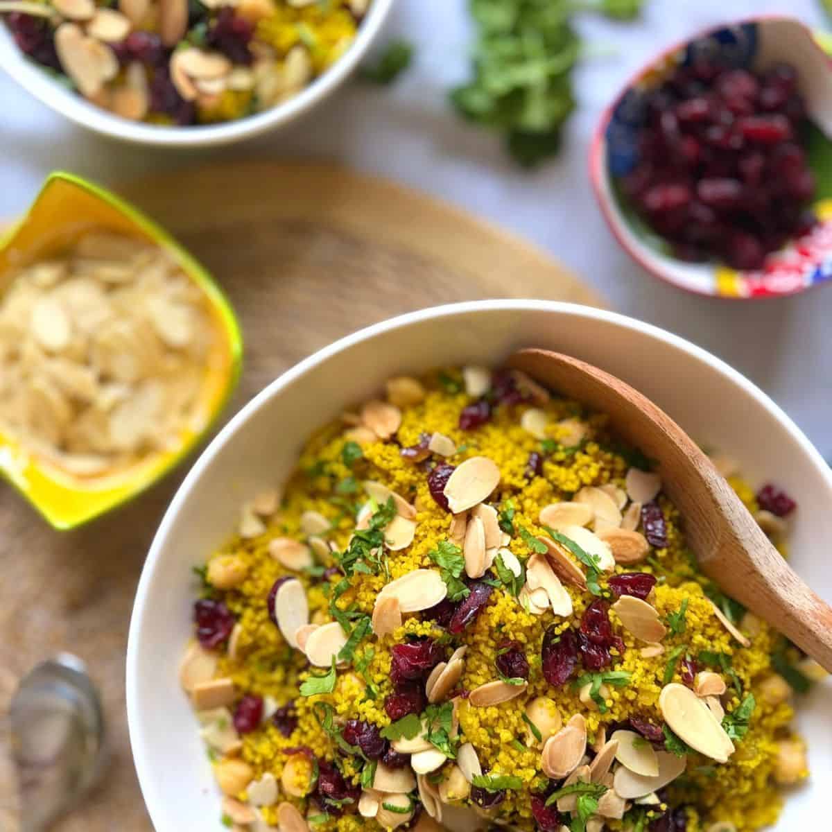 Morrocan couscous in a bowl on a table with a bowl of dried cranberries and a small bowl of flaked almonds